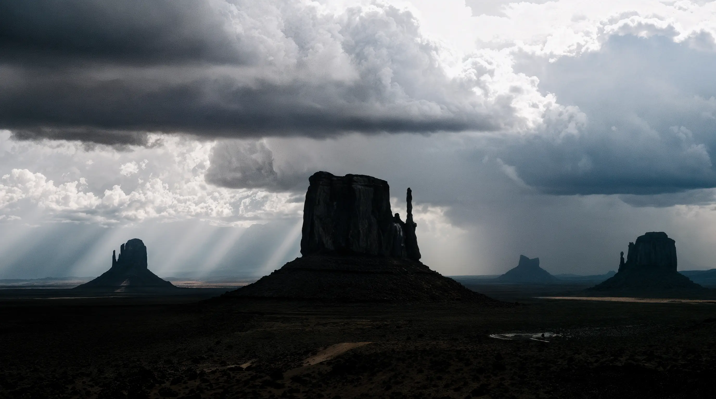 Dramatic desert landscape with towering buttes silhouetted against a stormy sky. Dark rain clouds loom overhead as sunbeams break through gaps, casting striking rays of light across the rugged terrain and highlighting the iconic rock formations